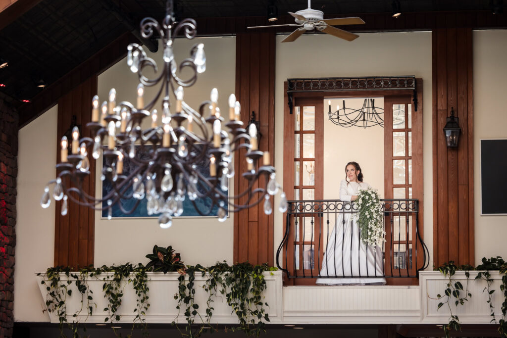 Bride stands gracefully on an indoor balcony at Fox Hollow, with a chandelier in the foreground and cascading bouquet in hand.