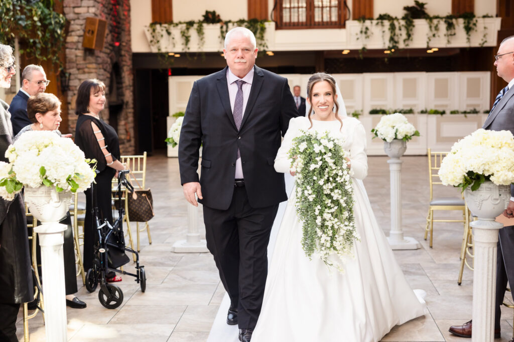 Bride walks down the aisle with her father, surrounded by smiling guests and lush white floral arrangements.
