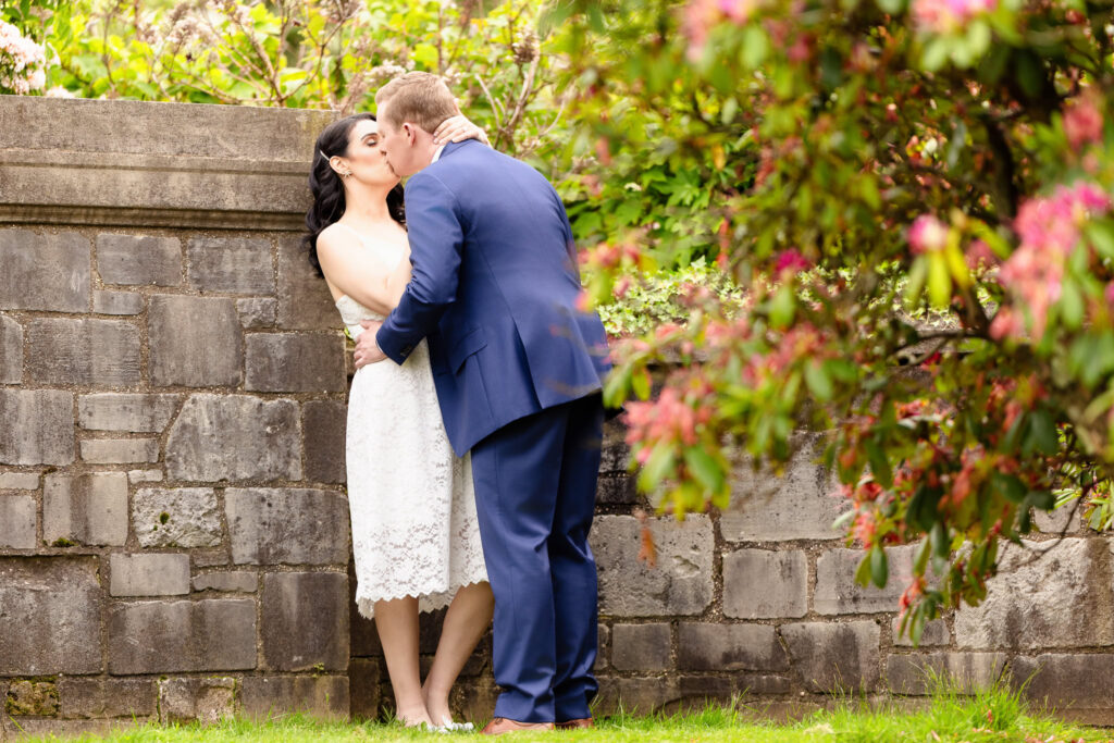 Romantic kiss between engaged couple Christine and Bill at Planting Fields Arboretum, framed by spring blossoms and stone garden walls in Long Island, NY
