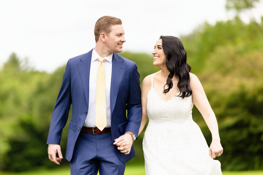 Christine and Bill smiling at each other while walking hand-in-hand through the gardens at Planting Fields Arboretum during their spring engagement session in Long Island