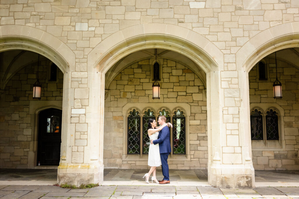 Christine and Bill embracing beneath a stone archway at Planting Fields Arboretum, framed by historic architecture and leaded glass windows during their elegant Long Island engagement session