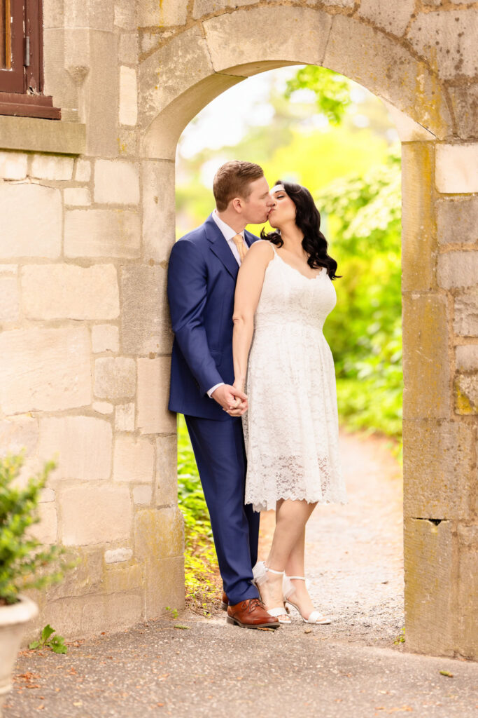 Christine and Bill sharing a kiss beneath a stone garden archway at Planting Fields Arboretum during their romantic Long Island engagement session