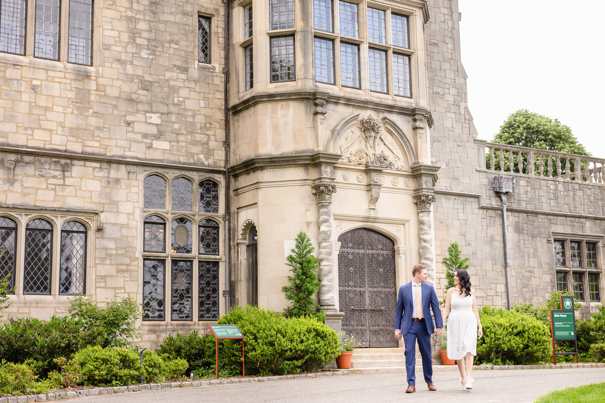 Engagement photo of a couple walking hand-in-hand in front of the grand stone facade of Planting Fields Arboretum's historic Main House in Long Island, NY