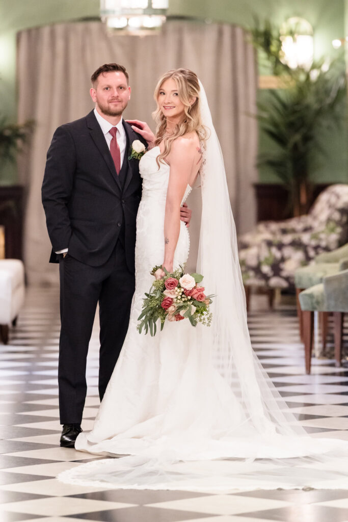 The bride and groom pose with quiet elegance in a grand interior space at The Heritage Club at Bethpage, her soft bouquet and flowing veil echoing the timeless beauty of the moment.
