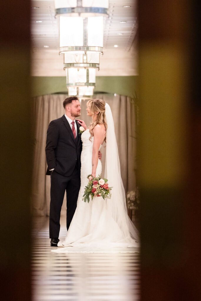 A romantic, softly lit portrait of the bride and groom captured through the doorway of The Heritage Club at Bethpage, highlighting the intimacy and elegance of their connection.
