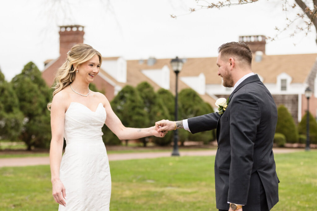 The bride and groom hold hands and smile during their first look on the grounds of The Heritage Club at Bethpage, capturing a tender and joyful start to their wedding day.