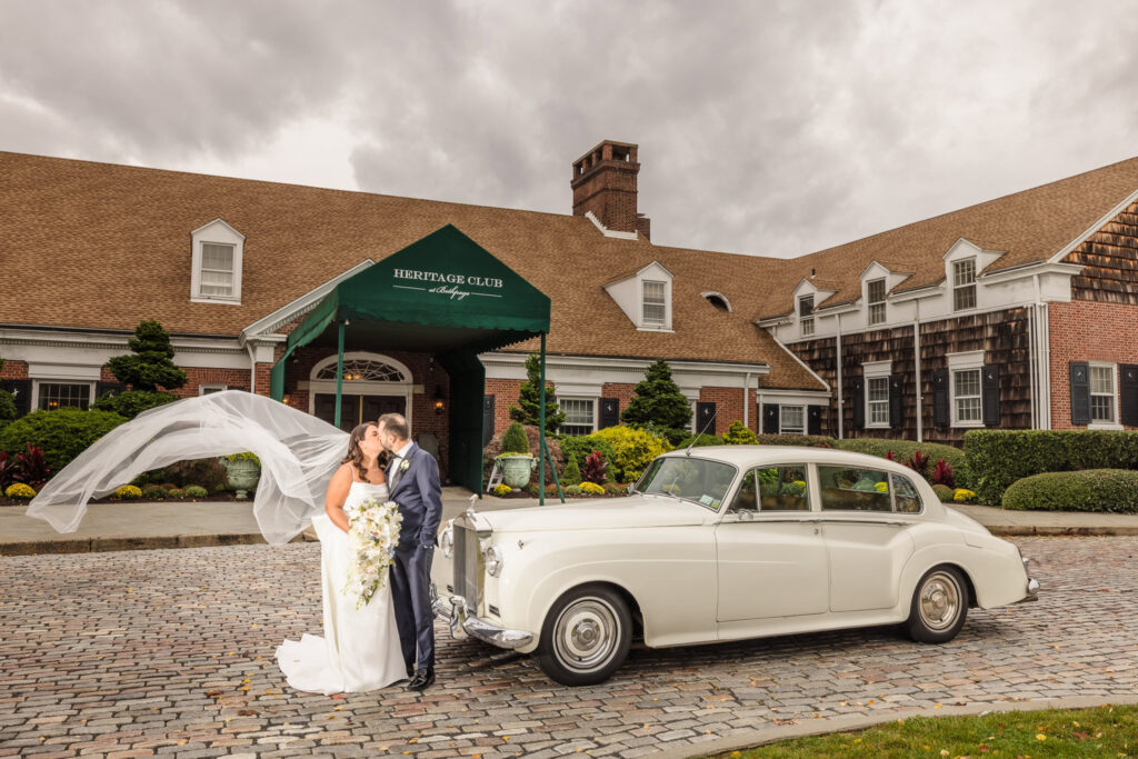 A newlywed couple shares a romantic kiss beside a vintage white car under the green awning of The Heritage Club at Bethpage, with the bride’s veil gracefully floating in the breeze—capturing timeless elegance and the beginning of their legacy.