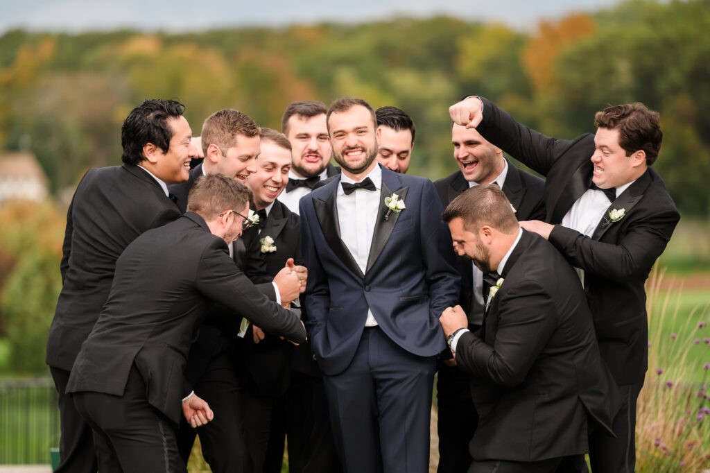 The groom shares a joyful, candid moment surrounded by his groomsmen on the grounds of The Heritage Club at Bethpage—laughter, brotherhood, and celebration before the vows.