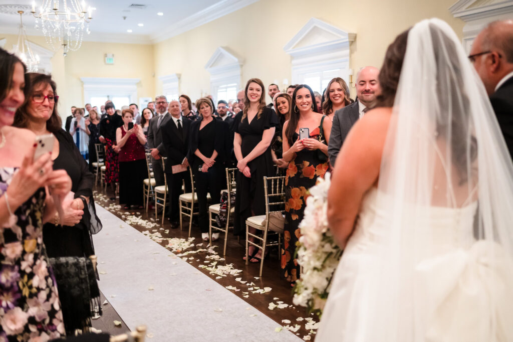 The bride walks down the aisle of the Lenox Room at The Heritage Club, surrounded by smiling guests and soft petals—an emotional, intimate beginning to the ceremony.