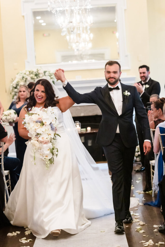 A bride and groom walk down the aisle at The Heritage Club at Bethpage. They are both holding their hands high and cheering. The bride is holding a cascading bouquet.