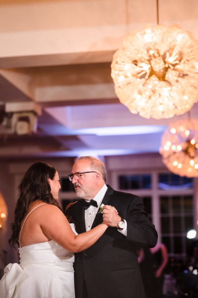 The bride and her father share a heartfelt dance under warm lighting at The Heritage Club, their connection and joy illuminated in a moment full of love and tradition.