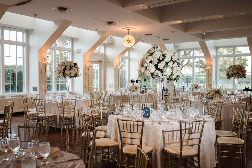 The Heritage Club at Bethpage Grand Ballroom set for a refined celebration, featuring gold chiavari chairs, ivory linens, and towering floral centerpieces bathed in natural light—an elegant and luminous space for timeless wedding memories.