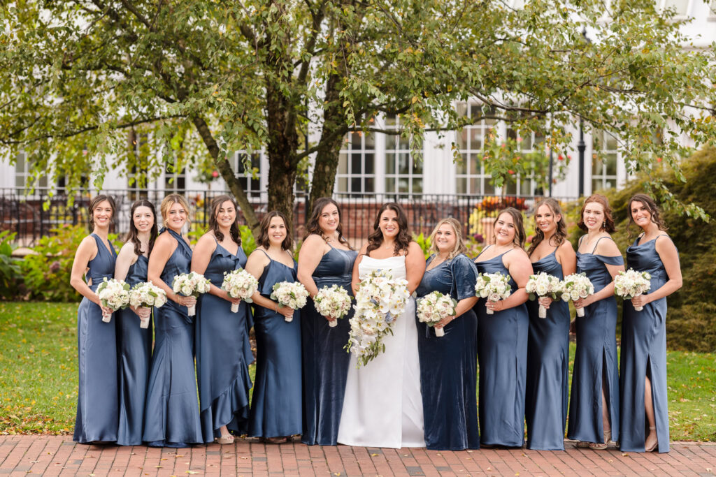 The bride poses with her bridesmaids dressed in elegant satin blue gowns, each holding white floral bouquets in front of the historic windows of The Heritage Club—radiating style, unity, and warmth.