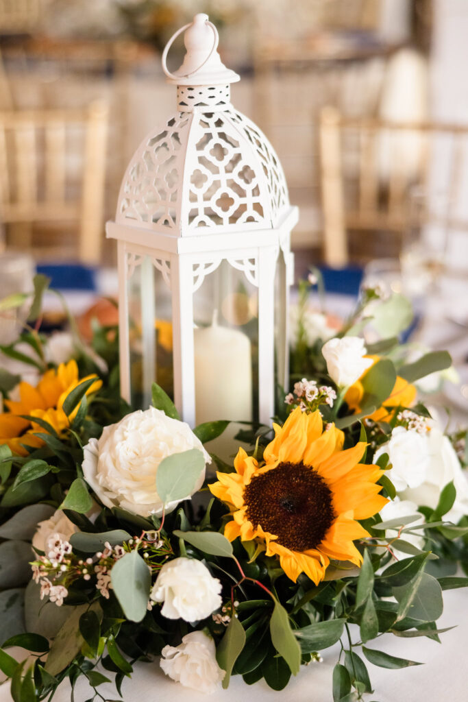 Close-up of a white lantern centerpiece surrounded by sunflowers, roses, and greenery on a reception table at Sea Cliff Manor.