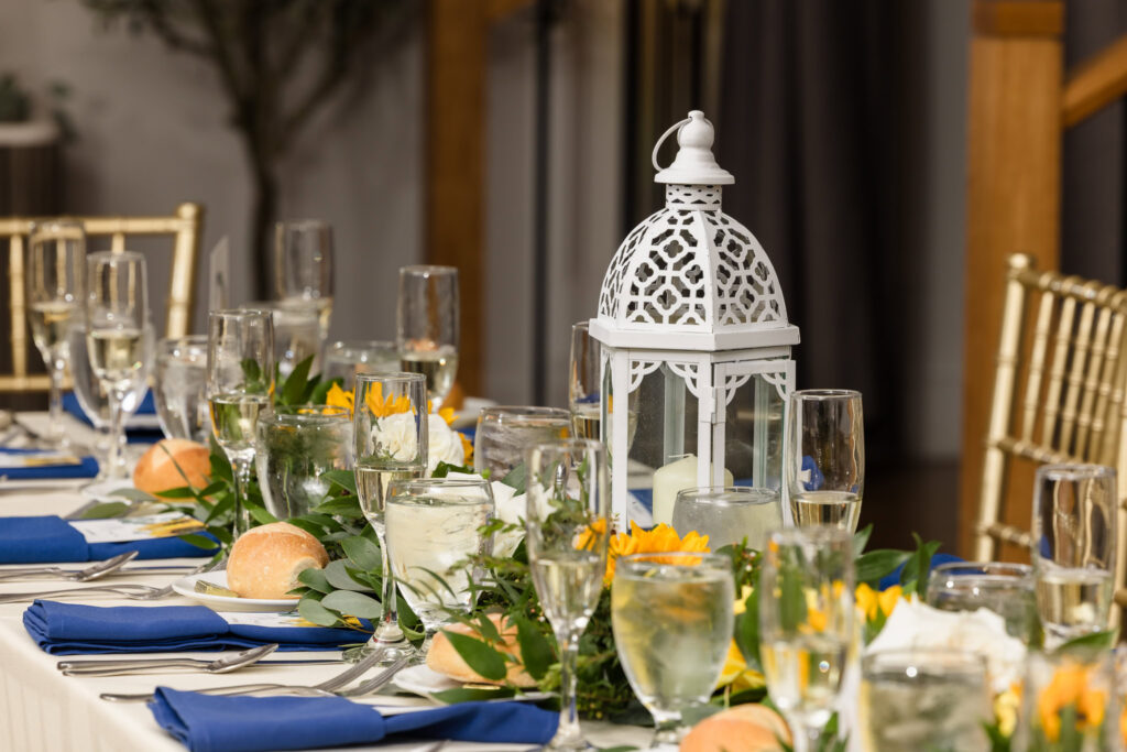 Close-up of an elegant reception table at Sea Cliff Manor featuring a white lantern centerpiece, greenery, yellow florals, and navy napkins.