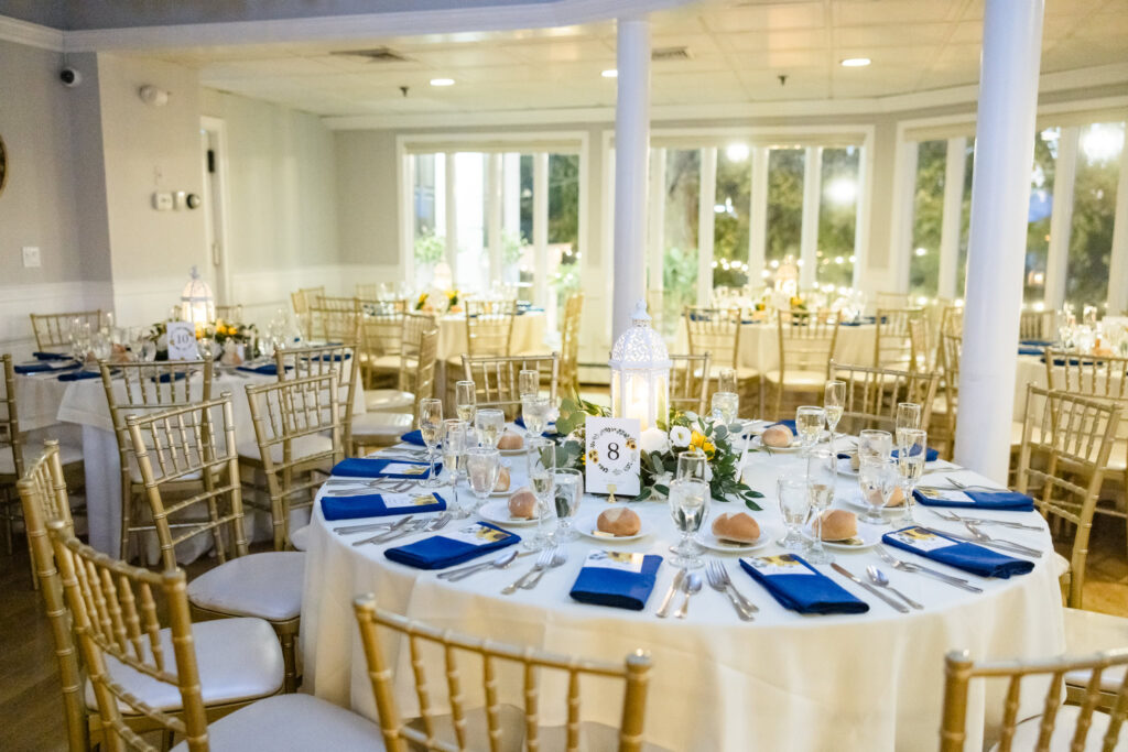 Reception table at Sea Cliff Manor set with gold chiavari chairs, white linens, navy napkins, and a romantic lantern centerpiece surrounded by florals and candlelight.
