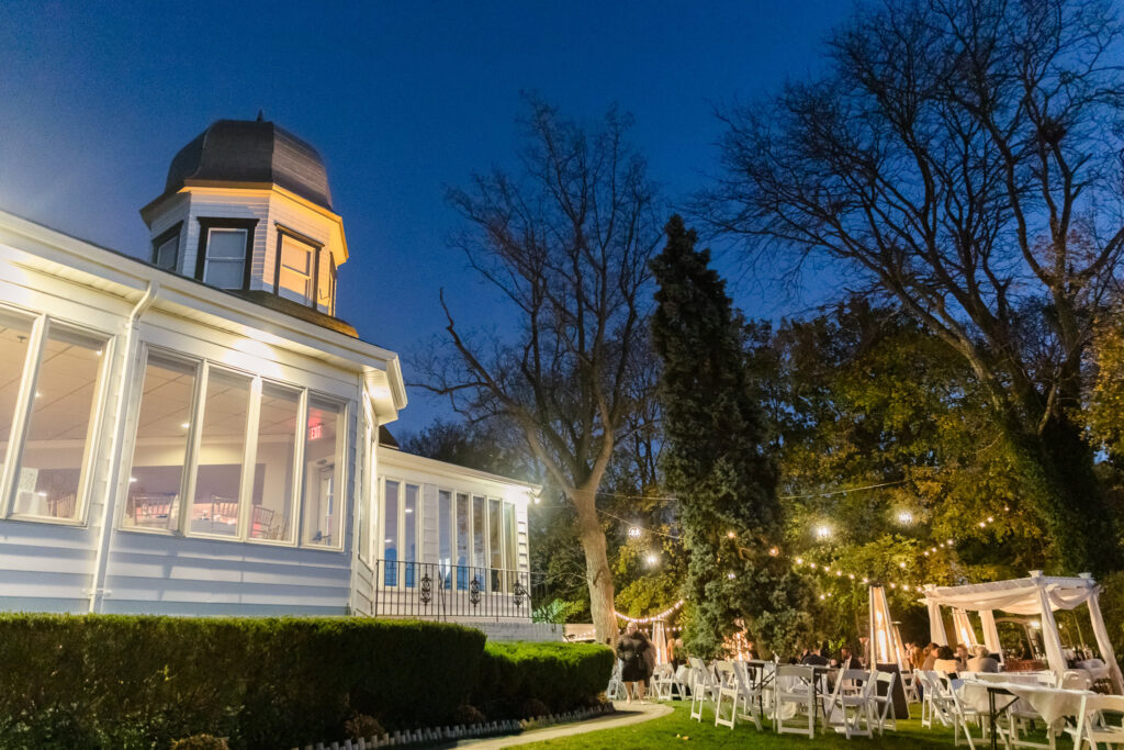 Twilight outdoor wedding reception at Sea Cliff Manor, with string lights, white seating, and the estate’s historic turret glowing under a deep blue sky.

