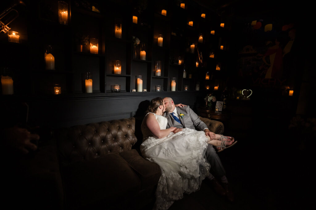 Bride and groom share an intimate kiss on a tufted leather sofa, surrounded by the warm glow of candlelight in the moody lounge at Sea Cliff Manor.