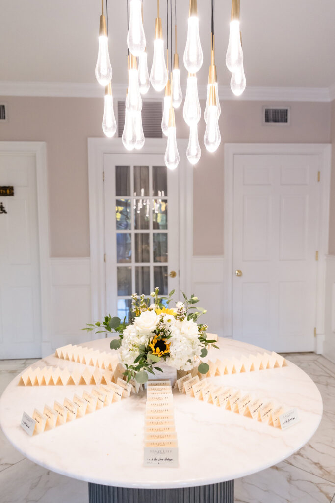 Round marble table with an escort card display, topped with fresh florals and lit by modern pendant lights in the entryway of Sea Cliff Manor.