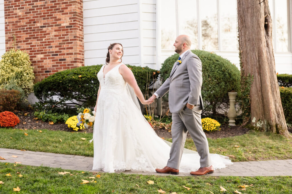 Bride and groom holding hands and smiling as they walk along the garden path at Sea Cliff Manor, framed by autumn blooms and historic architecture.