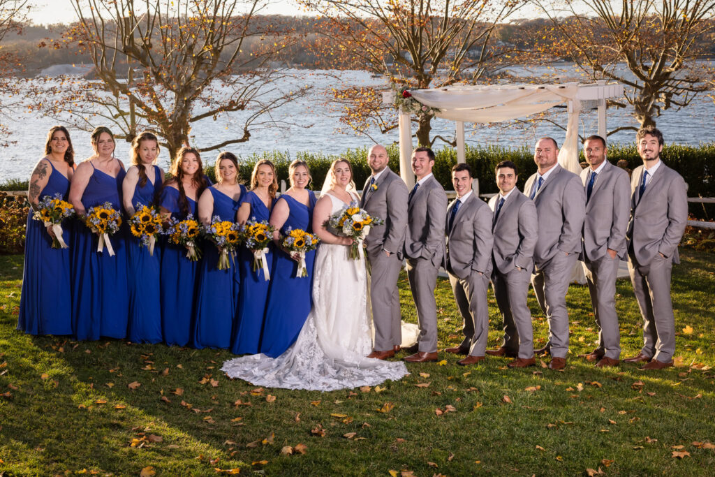 Bride and groom with their full wedding party in coordinated blue and gray attire, standing on the waterfront lawn at Sea Cliff Manor beneath a draped ceremony arbor.