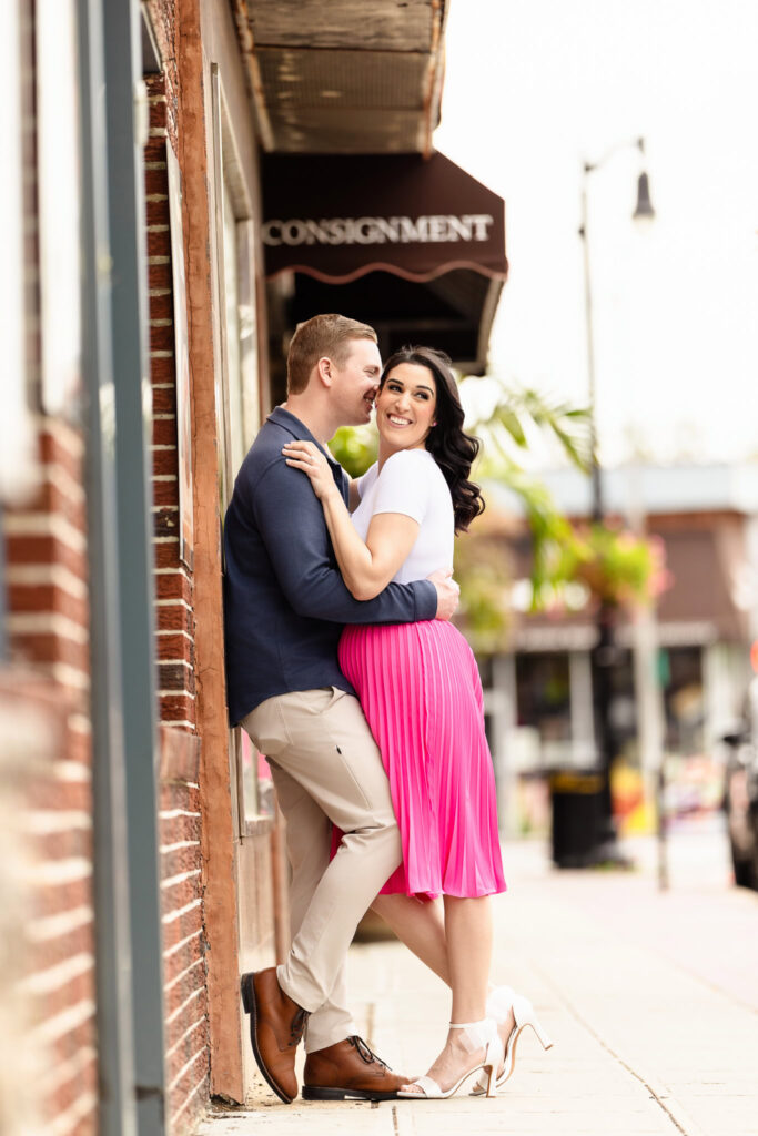 Christine and Bill sharing a sweet moment on the sidewalk outside a downtown Rockville Centre storefront during their engagement session, with warm smiles and a romantic embrace