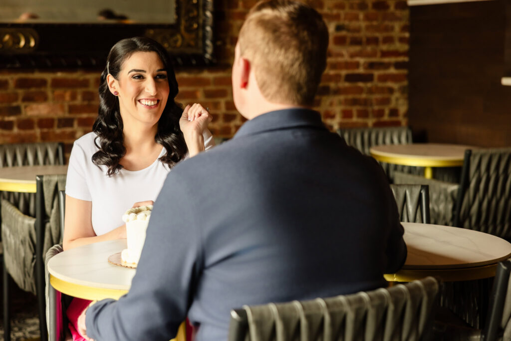 Christine smiling brightly at Bill during their engagement session inside Sugarberry café in Rockville Centre, seated across from each other with a small cake on the table