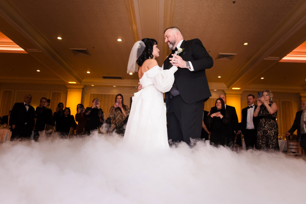 Bride and groom share their first dance surrounded by low-lying clouds on the ballroom floor at The Inn at New Hyde Park, as guests look on with joy.

