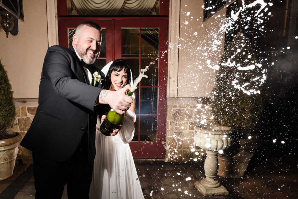 Bride and groom pop a bottle of champagne outside The Inn at New Hyde Park, laughing joyfully as bubbles and spray capture the excitement of their celebration.


