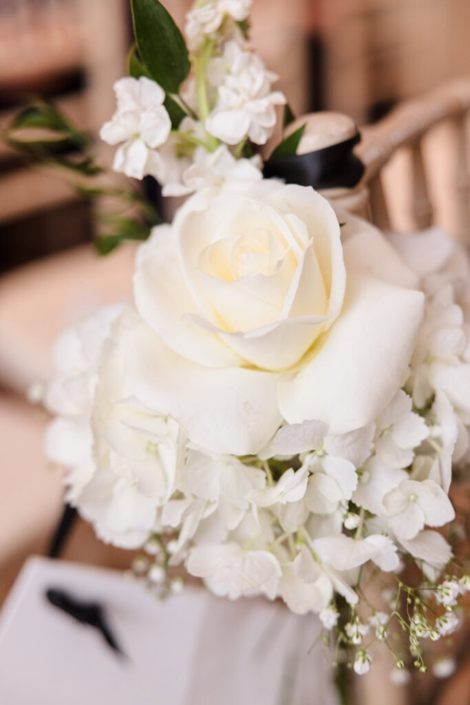 Close-up of a white rose and hydrangea floral arrangement tied to a ceremony chair with black ribbon at The Inn at New Hyde Park.

