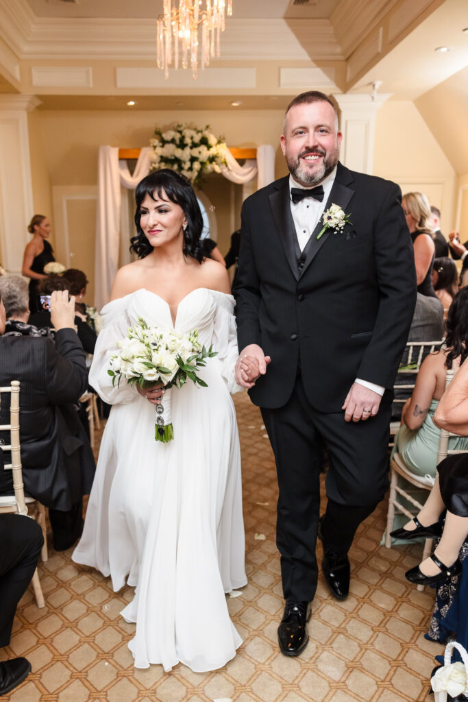 Joyful bride and groom walk hand-in-hand down the aisle at The Inn at New Hyde Park, just moments after saying “I do,” surrounded by loved ones and soft, elegant decor.

