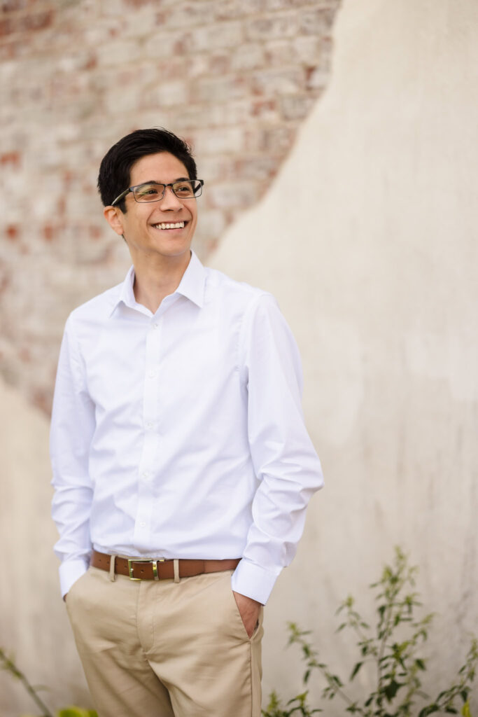 Groom-to-be standing relaxed against a textured wall with a warm smile, dressed in classic attire for their engagement session.
