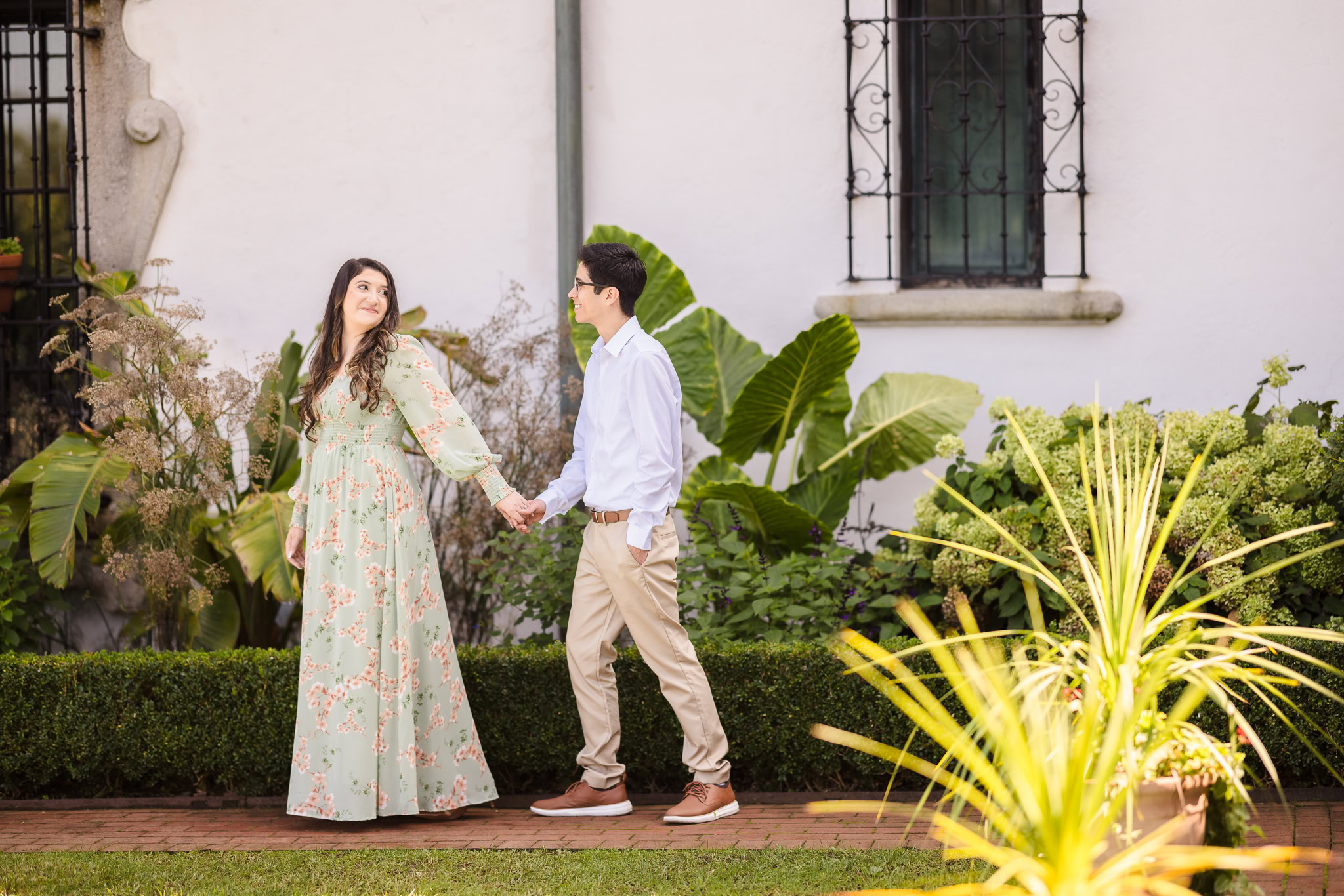 Couple holding hands and walking through the Vanderbilt Museum gardens, surrounded by lush greenery and warm afternoon light during their fall engagement session.