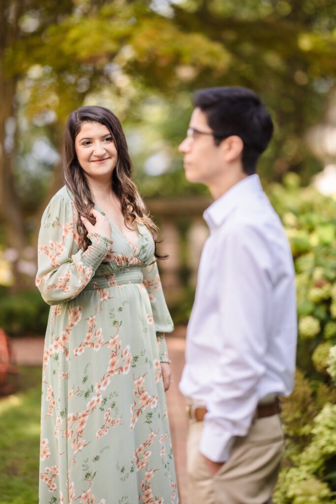 Bride-to-be softly smiling at the camera while her fiancé gazes at her, surrounded by garden greenery at the Vanderbilt Museum.