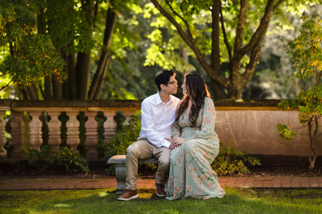 Couple sitting closely on a stone bench in the Vanderbilt Museum gardens, sharing an intimate gaze beneath the dappled light of surrounding trees.