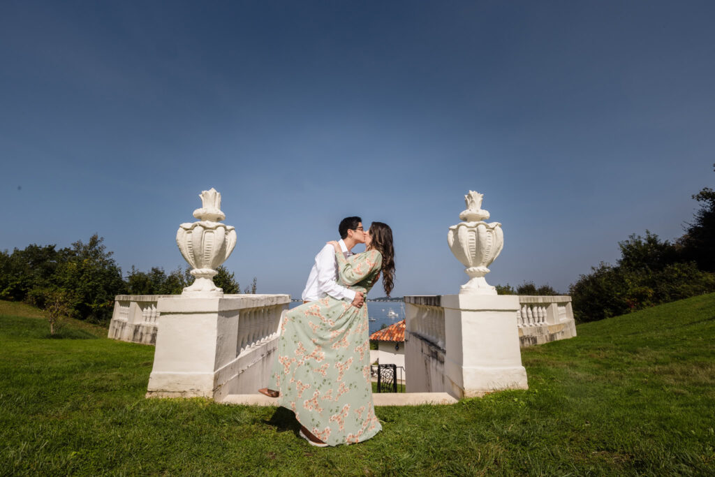 Groom-to-be lifting his fiancée in a joyful kiss on the grand terrace steps of the Vanderbilt Museum, with sweeping coastal views under a clear autumn sky.