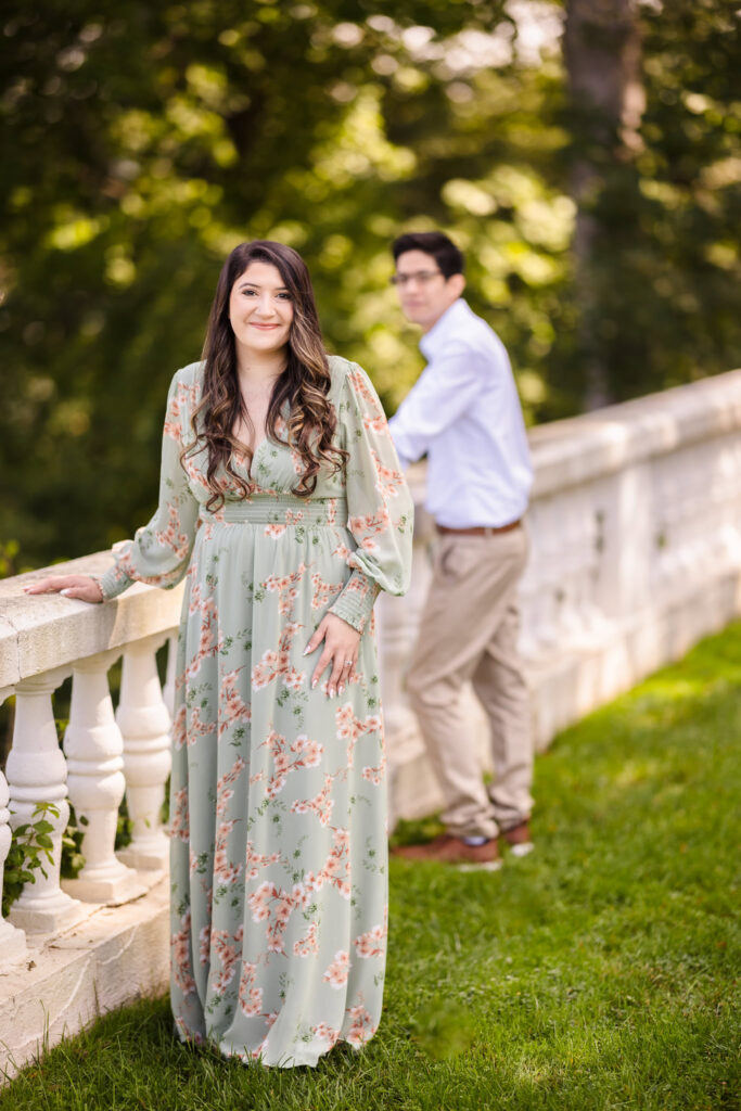 Bride-to-be standing gracefully along the balustrade at the Vanderbilt Museum, with her fiancé walking softly in the background.