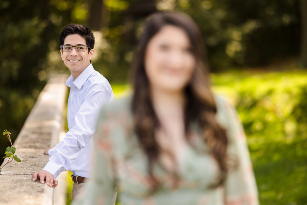 Groom-to-be smiling softly while leaning on a stone ledge at the Vanderbilt Museum, with his fiancée blurred in the foreground, creating a romantic depth of field.