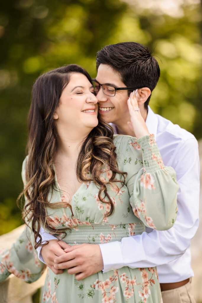 Couple embracing with joy during their engagement session, as he gently kisses her cheek against a sun-dappled, leafy backdrop.