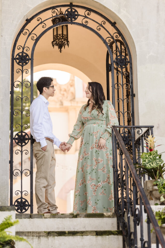 Couple holding hands and smiling at each other beneath the ornate arched iron gate at the Vanderbilt Museum.