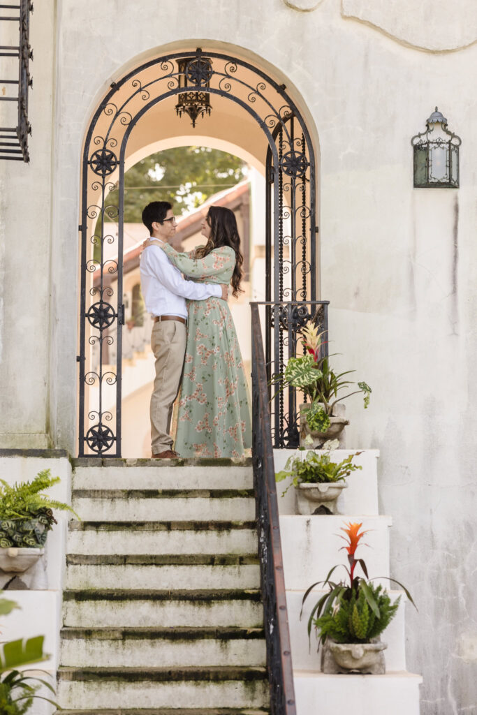 Couple embracing under the grand iron arch at the top of the staircase, surrounded by soft light and architectural charm.