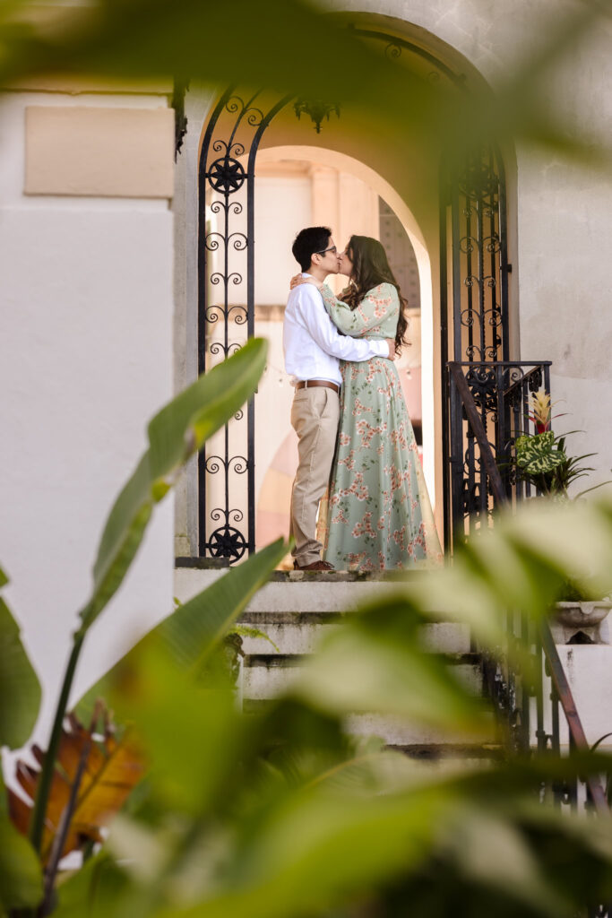  Intimate embrace partially hidden behind lush garden leaves, with the couple framed in the museum’s elegant arch.