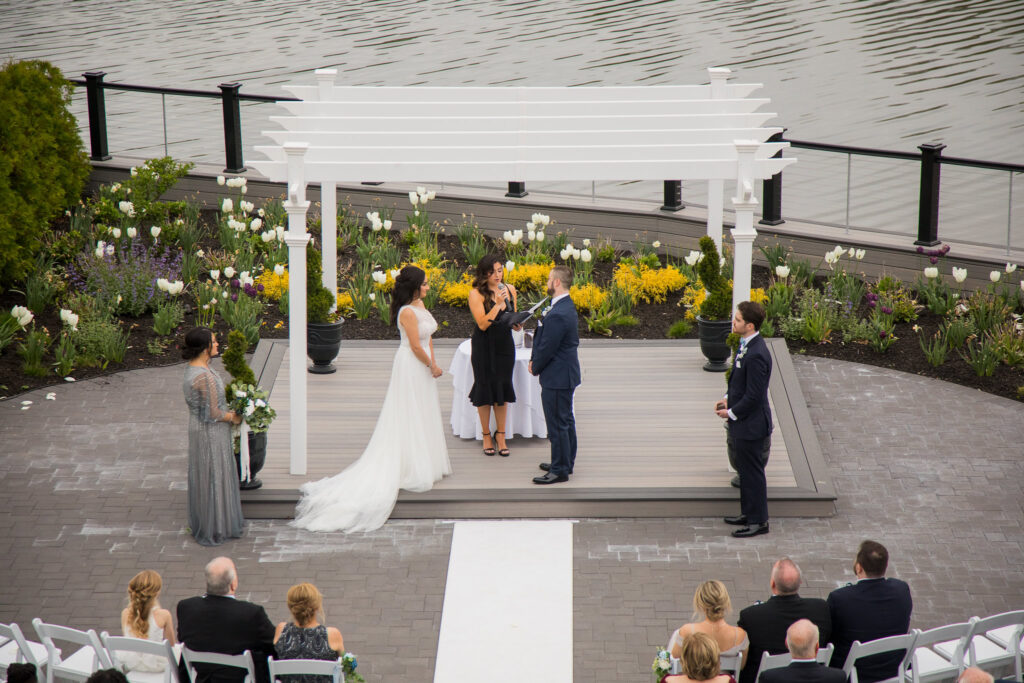 Waterfront wedding ceremony under a white pergola surrounded by spring flowers at Water’s Edge Centerport.
