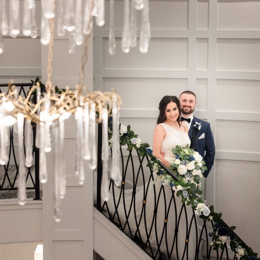 Bride and groom pose on elegant staircase adorned with florals and chandelier at Water’s Edge Centerport.