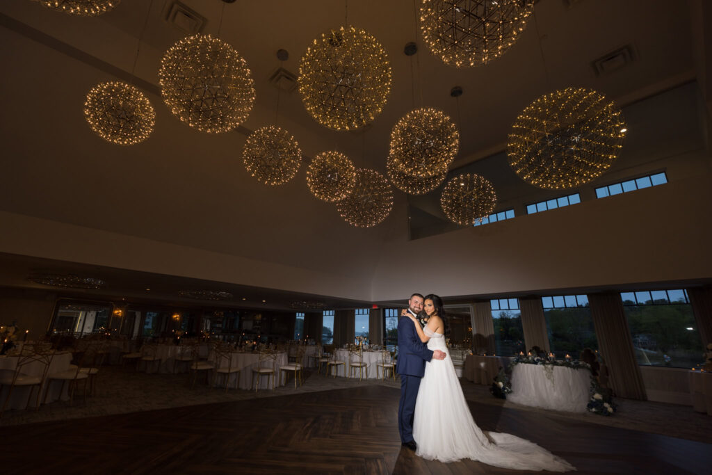 Bride and groom share a romantic moment under glowing chandeliers in the ballroom at Water’s Edge Centerport.