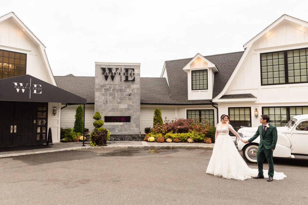 Bride and groom hold hands in front of Water’s Edge Centerport with classic white car and venue facade in background.