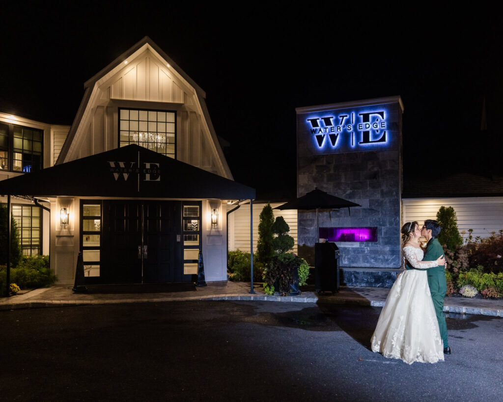 Bride and groom share a romantic nighttime moment in front of the illuminated Water’s Edge Centerport entrance.