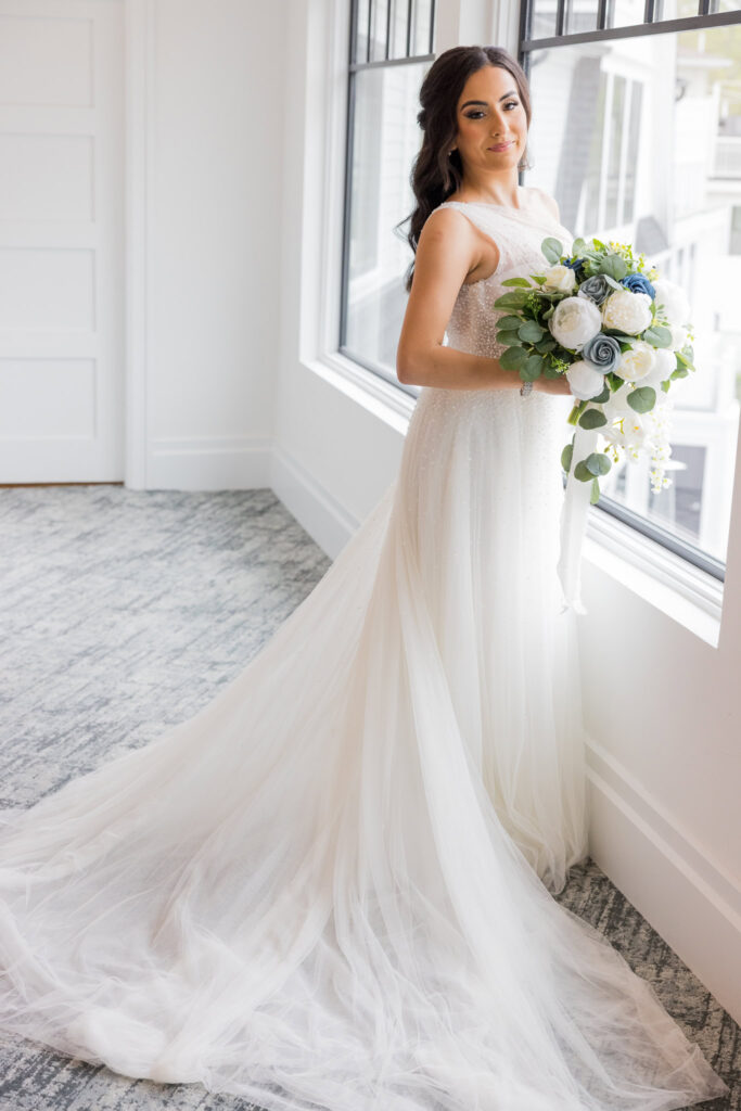 Bride poses with bouquet in front of large window, showcasing her gown at Water’s Edge Centerport.