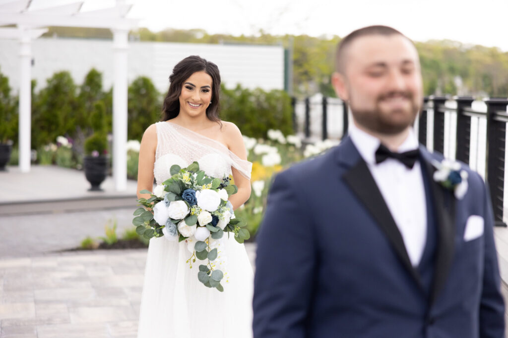 Bride approaches groom from behind with a smile during their first look at Water’s Edge Centerport.