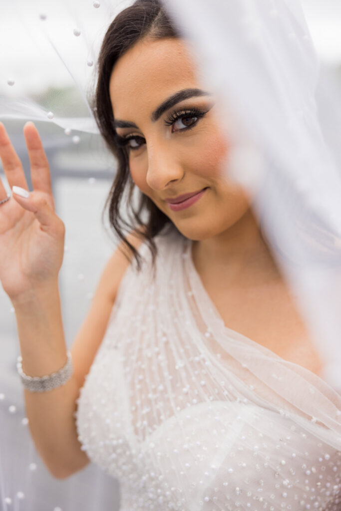 Bride softly lifts her veil while smiling during a quiet moment at Water’s Edge Centerport.